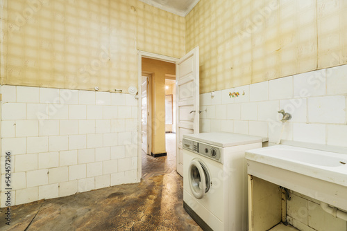 Cocina vieja, sucia y destartalada con suelo oscuro, azulejos blancos y papel amarillo en las paredes photo