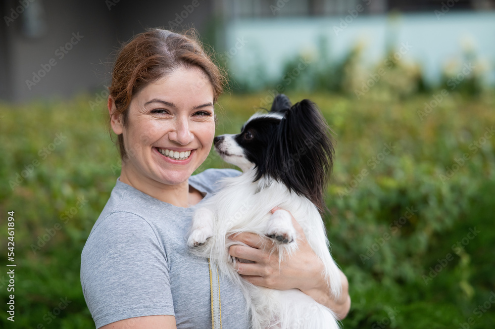 Caucasian red-haired woman holding pappilion dog in her arms outdoors. Black and white continental spaniel. Caucasian red-haired woman holding pappilion dog in her arms outdoors. Black and white continental spaniel.