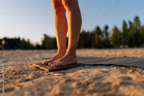 Close-up feet of unrecognizable woman standing barefoot on wooden Sadhu nail board during meditation practice on sandy beach. Closeup cropped shot of female practicing yoga on fire board at fresh air.