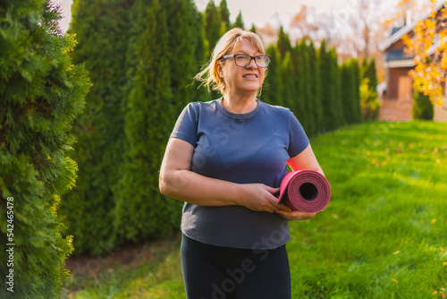 Standing and holding yoga mat. Senior woman having nice weekend outdoors on the field at sunny day.