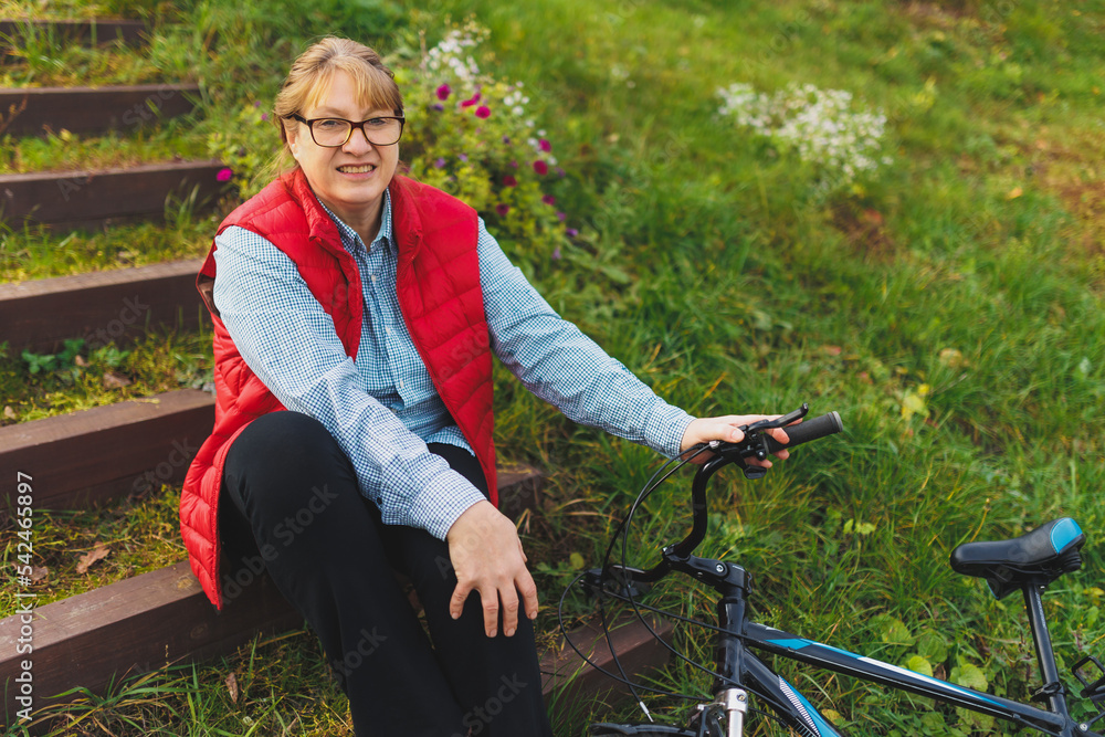 Fototapeta premium Middle aged smiling mature woman holdingt a bike with her hands on the grass on a green field. Summer or Autumn Country Vacation and Adventure Concept