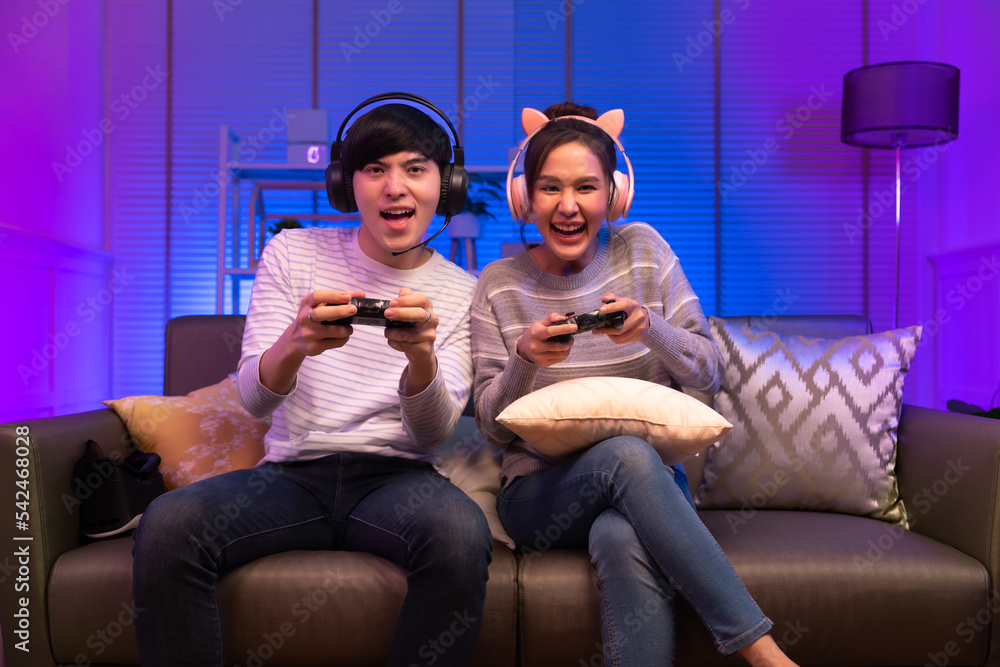 Young attractive asian woman playing video games at home neon lighted living room . Side view of a couple working in front of the same workstation.
