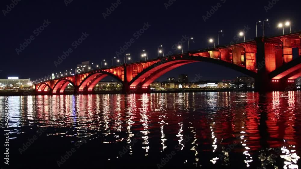 Night landscape with red illumination of Communal Bridge in Krasnoyarsk city, Russia. View of the Yenisey River at night. Reflection of lighting lights in the rippled water surface Night landscape with red illumination of Communal Bridge in Krasnoyarsk city, Russia. View of the Yenisey River at night. Reflection of lighting lights in the rippled water surface