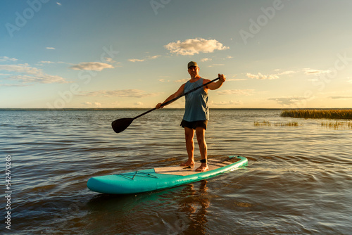 A man in shorts on a SUP board with a paddle floats on the water in the rays of the setting sun.
