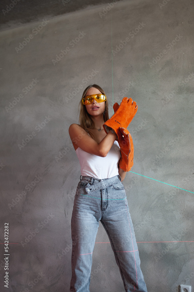 Beautiful blonde girl stands with construction tools in construction uniform in orange gloves and yellow glasses