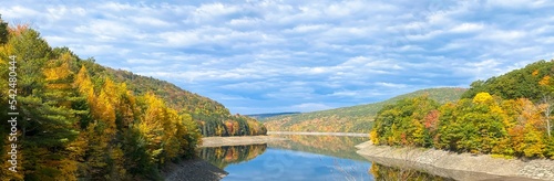 Fototapeta Naklejka Na Ścianę i Meble -  Calm lake surrounded by forested hills with
vibrant autumn leaf color; reflections in water, low
water levels, blue sky with clouds. Pepacton Reservoir, New York State, US