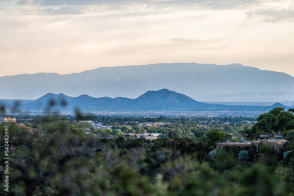 Naklejka premium Colorful sunset in Santa Fe, New Mexico skyline with green foliage in foreground and cityscape buildings with mountains silhouette