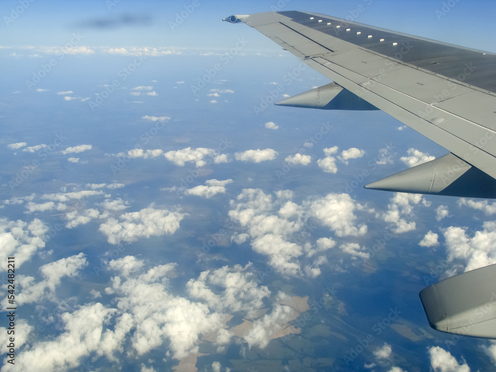 The wing of an airplane in the blue sky. There are cumulus clouds under it and the earth in the distance. The wing of an airplane in the blue sky. There are cumulus clouds under it and the earth in the distance.