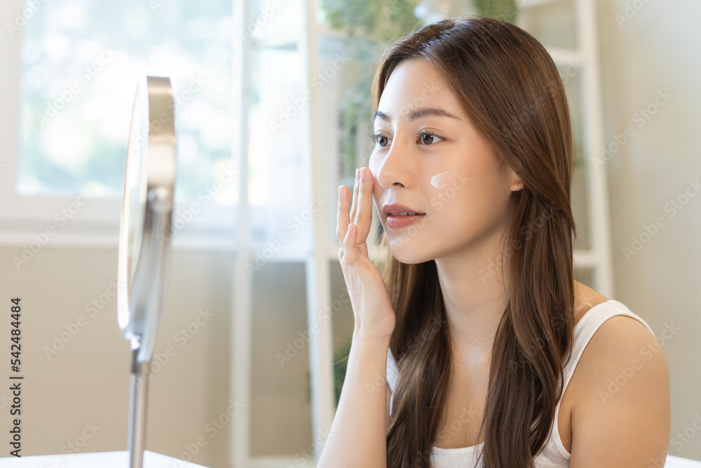 Fototapeta premium Fresh healthy skin, beautiful smile of asian young woman, girl looking at mirror, applying moisturizer on her face, putting cream treatment before make up cosmetic routine at home. Facial Beauty.