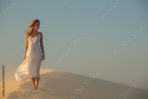 A girl in a white dress fluttering in the wind walks through the desert in the sunset light.