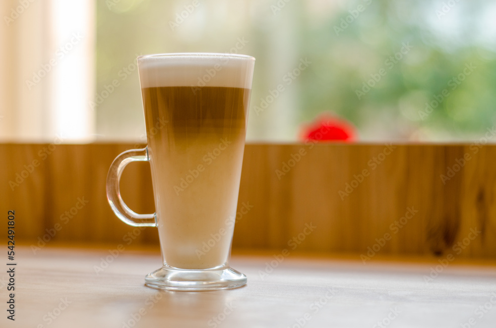 Beautiful tall transparent glass with a handle containing a latte coffee drink on a table, in a cafe in front of a window with natural light Beautiful tall transparent glass with a handle containing a latte coffee drink on a table, in a cafe in front of a window with natural light
