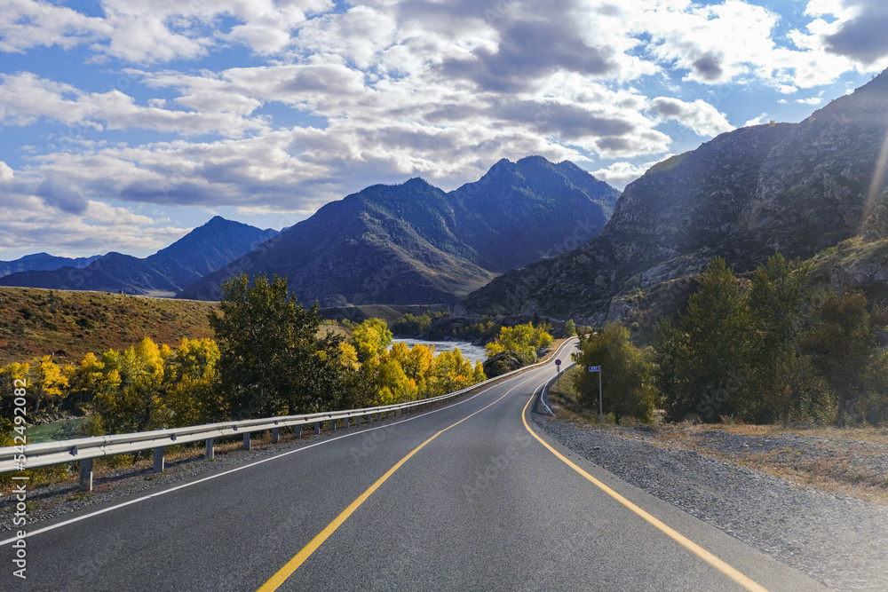 Fototapeta premium Serpentine asphalt road among high snow-capped mountain peaks, yellow desert, autumn green forest and blue sky. Car on the highway against the background of a mount landscape.