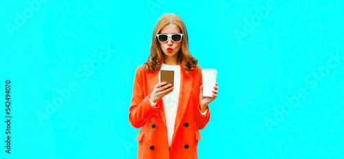 Portrait of beautiful young woman with smartphone wearing red jacket on blue background