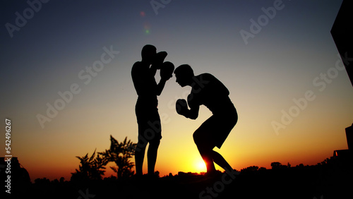 Two dark male figures  at sunrise  against the light  boxing  fighting in sparring  training in a pair of techniques of strikes. On sandy beach  in cargo port  near water  in summer. High quality