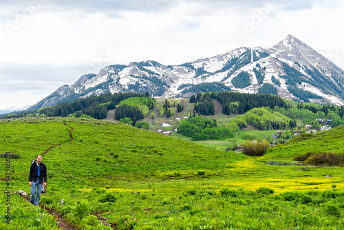 Happy young man standing on Snodgrass trail hike with camera and view of Mount Crested Butte, Colorado peak and yellow buttercup wildflowers photo