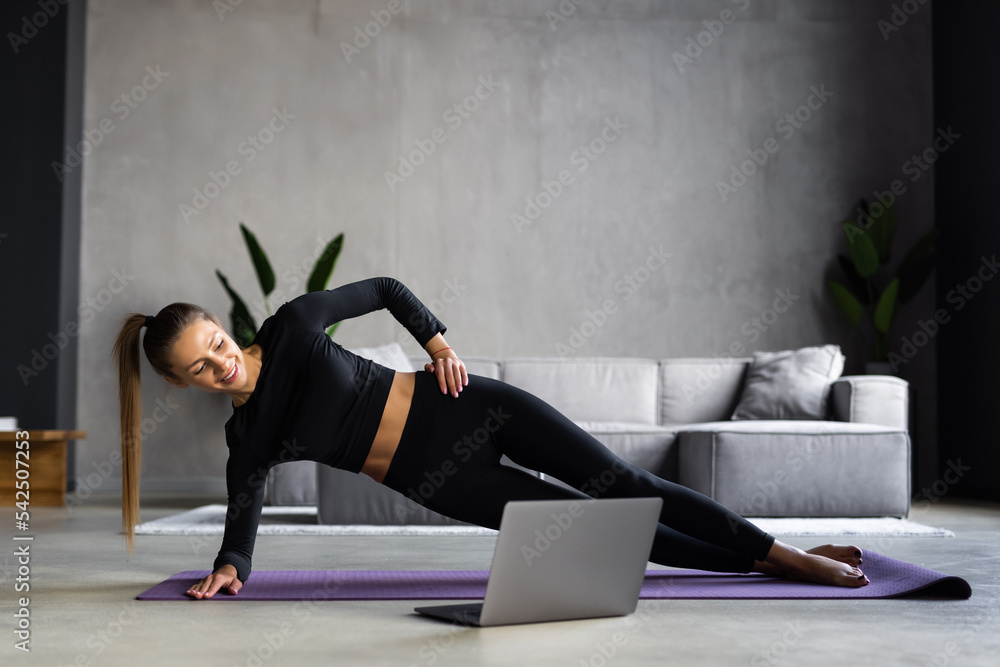 Beautiful sporty cheerful woman is doing side plank exercise watching video on the internet and repeating the tasks