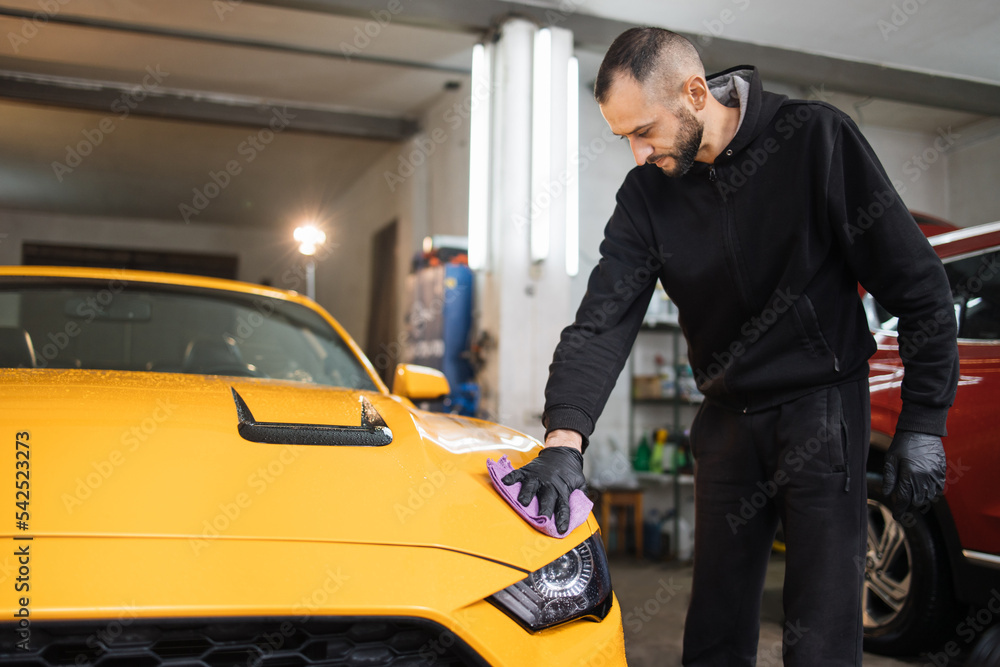Car detailing and cleaning concept. Male car wash worker holds the microfiber in hand and polishes the car hood. Selective focus on the hand with microfiber. Car detailing and cleaning concept. Male car wash worker holds the microfiber in hand and polishes the car hood. Selective focus on the hand with microfiber.