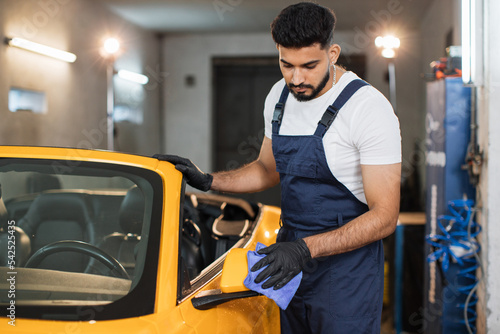 Bearded male car service worker in black rubber glove and overalls holding the microfiber cloth and cleaning car mirror of back view at modern car wash service.