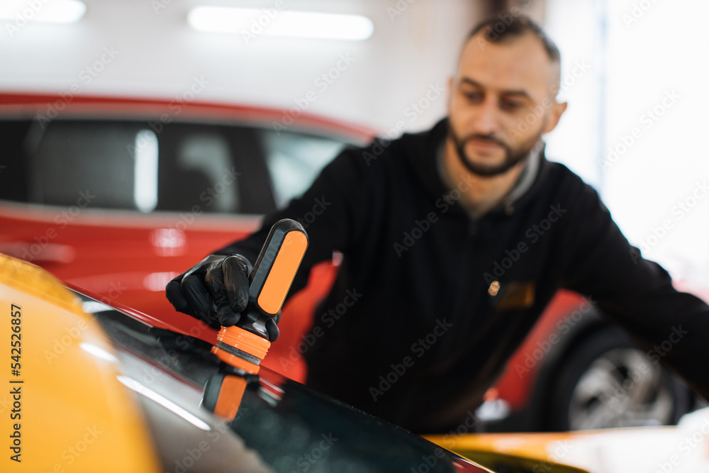 Focus on hand of bearded male worker in black uniform and protective gloves, on a professional car wash service, applying anti rain coating on a windshield of luxury yellow car. Focus on hand of bearded male worker in black uniform and protective gloves, on a professional car wash service, applying anti rain coating on a windshield of luxury yellow car.