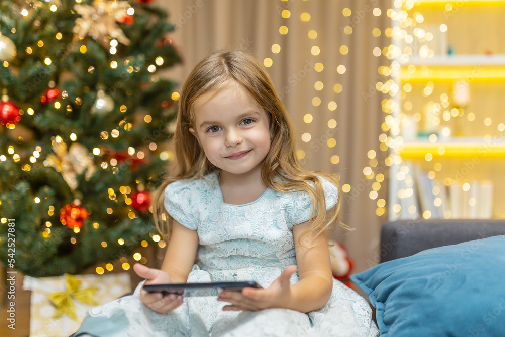 Portrait of a little girl at home with a smartphone sitting on the sofa near the Christmas tree watching video content online on Christmas and New Year's holidays, looking at the camera and smiling. Portrait of a little girl at home with a smartphone sitting on the sofa near the Christmas tree watching video content online on Christmas and New Year's holidays, looking at the camera and smiling.