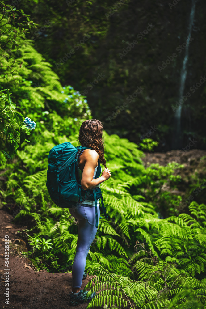 Sporty woman marvels at an impressive waterfall in the jungle with beautiful flowers and ferns. Levada of Caldeirão Verde, Madeira Island, Portugal, Europe.