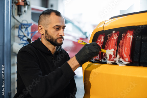 Close up photo of young male worker in black protective gloves cleaning tail light in foam with special brush. Sport yellow luxury car wash and detailing. Washing machine at the station. © sofiko14