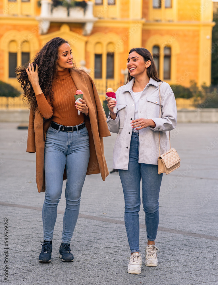 Two girls are strolling, talking and eating cone ice cream, and having much fun in the town.