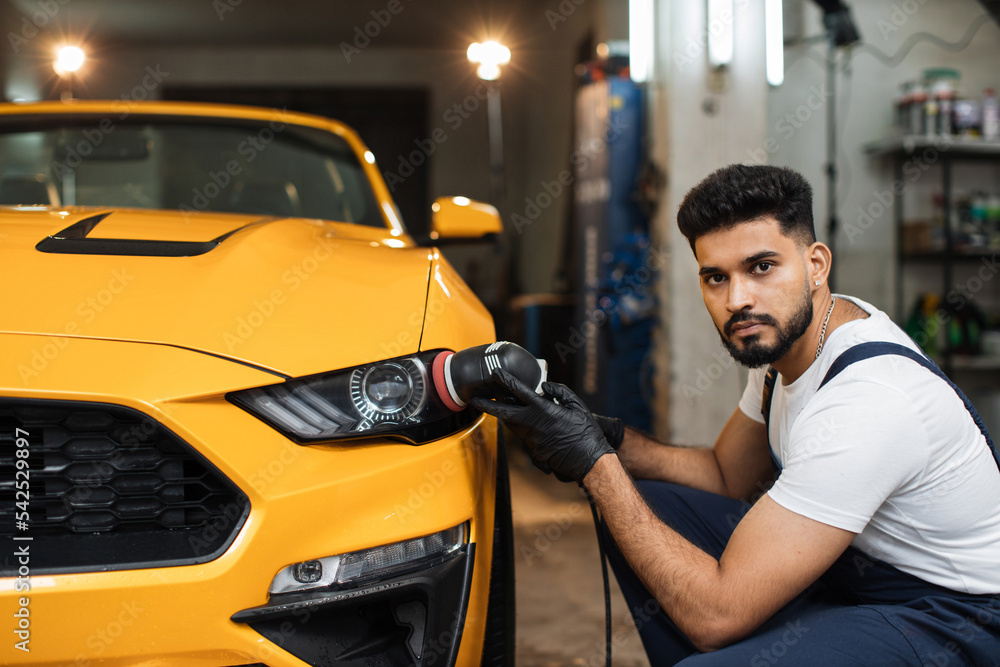Young professional bearded male worker in overalls and white t-shirt with orbital polisher in auto service polishing headlights of yellow luxury car. Detailing and polishing of car headlights. Young professional bearded male worker in overalls and white t-shirt with orbital polisher in auto service polishing headlights of yellow luxury car. Detailing and polishing of car headlights.