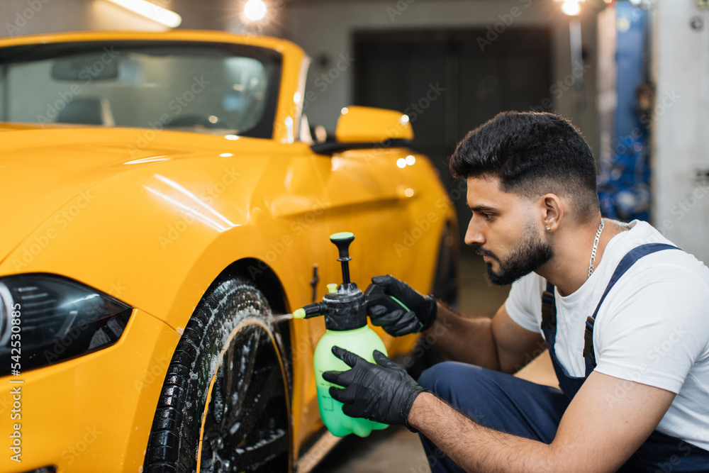 Washing a car in detailing service. Indoor shot of male worker in overalls, washing the car wheels rims with pressure water sprayer after cleaning it with special cleansing solution. Washing a car in detailing service. Indoor shot of male worker in overalls, washing the car wheels rims with pressure water sprayer after cleaning it with special cleansing solution.