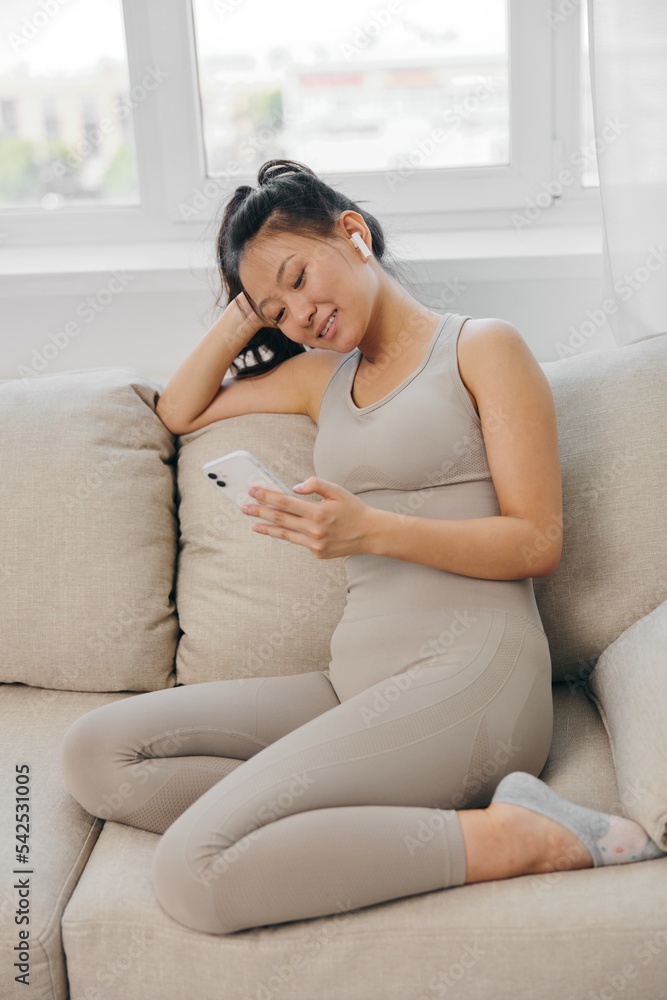 Athletic Asian woman in yoga suit blogger looking at phone in hand sitting on couch and smiling with headphones, freelance work from home, recording sports workout lifestyle body care Athletic Asian woman in yoga suit blogger looking at phone in hand sitting on couch and smiling with headphones, freelance work from home, recording sports workout lifestyle body care