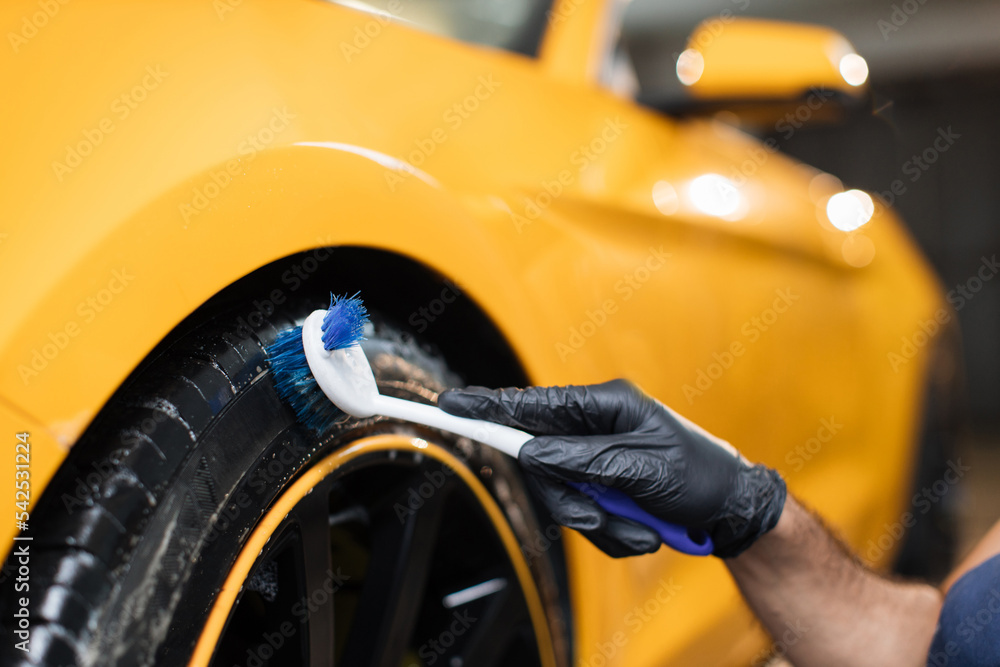 Auto wash service. Cropped close up image of male hands in black protective gloves, cleaning wheels tires of luxury car with a special brush in a vehicle detailing workshop.