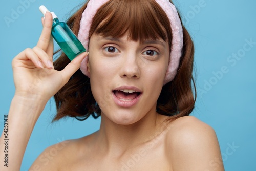 horizontal beauty photo on a blue background of a pleasant woman with clean skin standing on a blue background and holding a jar of skin serum near her face, her mouth wide open with delight