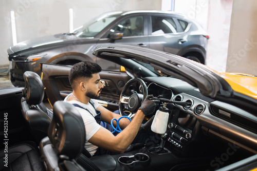 Handsome male cleaner in overalls, white t-shirt and protective gloves, removing dirt from car salon using professional aero chemical cleaner gun.