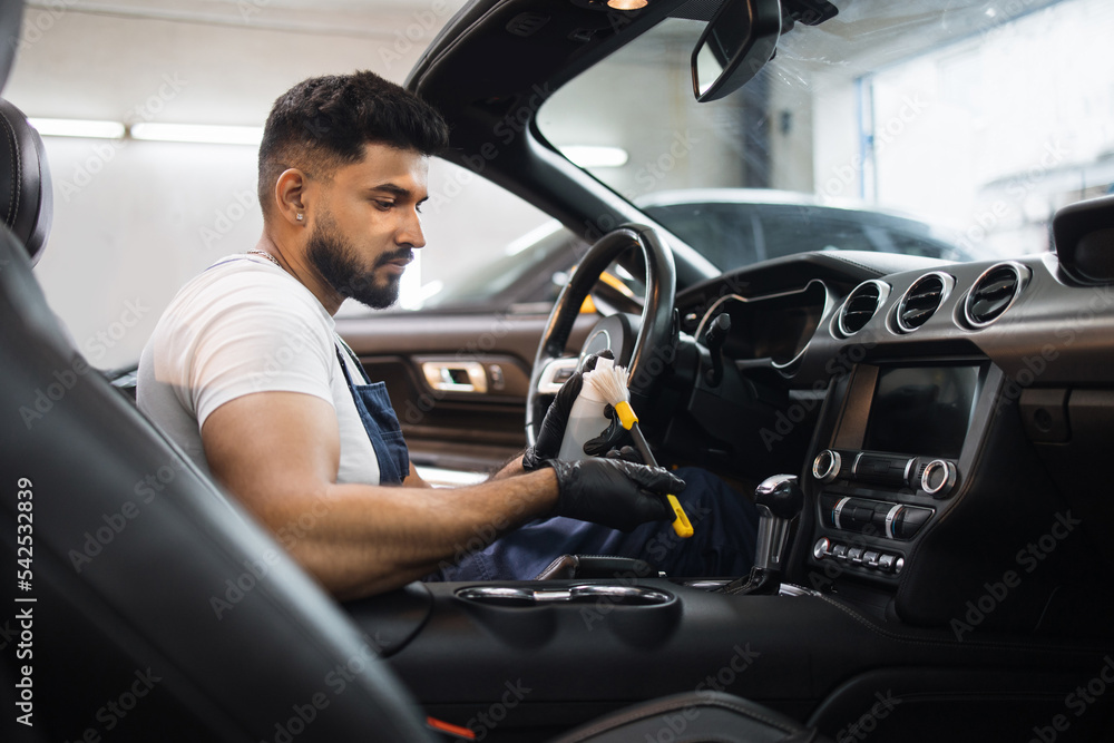 Bearded young male car detailing worker in overalls, sitting inside of modern vehicle, and cleaning car dashboard, putting plastic trim care milk on the soft brush