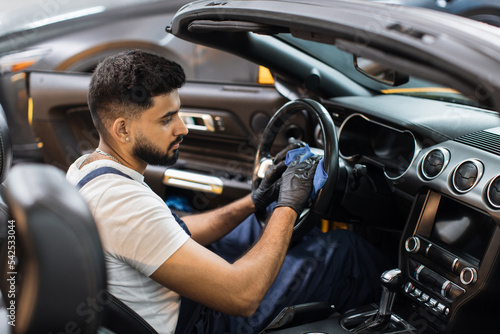 Handsome bearded young man in uniform and protective gloves, cleaning car interior and steering wheel using microfiber cloth, smiling at camera. Car detailing and valeting concept.