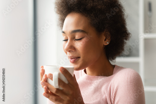 Happy pretty young african american female with curly hair, with closed eyes enjoy favorite drink in cup