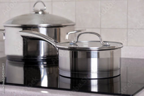 Ladle and large saucepan stand on modern stove with a glass-ceramic hob. Stainless steel utensils. Professional and home kitchen equipment and utensils. Selective focus, copy space.