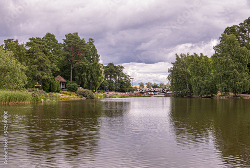 Finland, Kotka - July 18, 2022: Sopokanlahti park and lake. Whtie wooden bridge spans water link to harbor and sea under gray cloudscape. Green tree belt on both sides photo