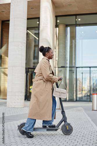 Vertical side view portrait of young black woman riding electric scooter in city and smiling cheerfully while wearing long trenchcoat