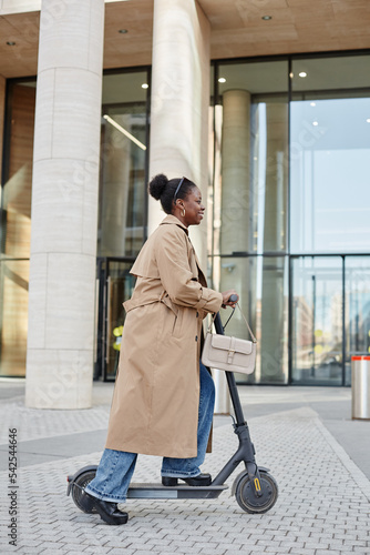 Vertical side view portrait of young black woman riding electric scooter in city and smiling cheerfully while wearing long trenchcoat photo