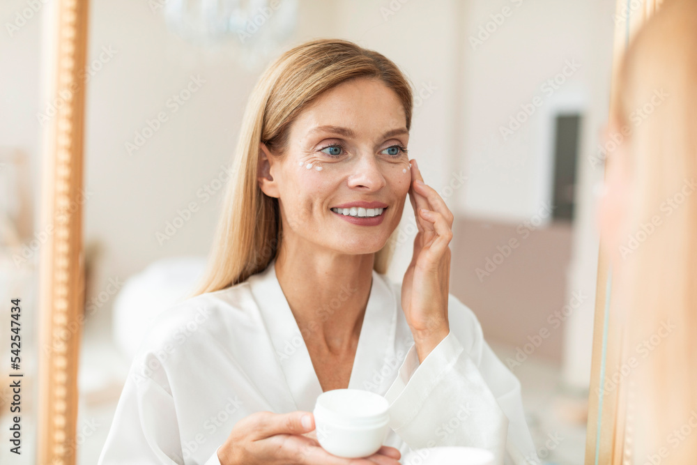 Happy blonde middle aged woman in bathrobe using moisturizing eye cream, looking at mirror, enjoying face care routine Happy blonde middle aged woman in bathrobe using moisturizing eye cream, looking at mirror, enjoying face care routine