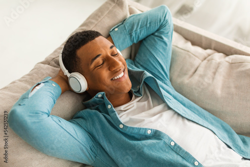 Satisfied millennial african american male lies on sofa in wireless headphones with closed eyes, enjoys music