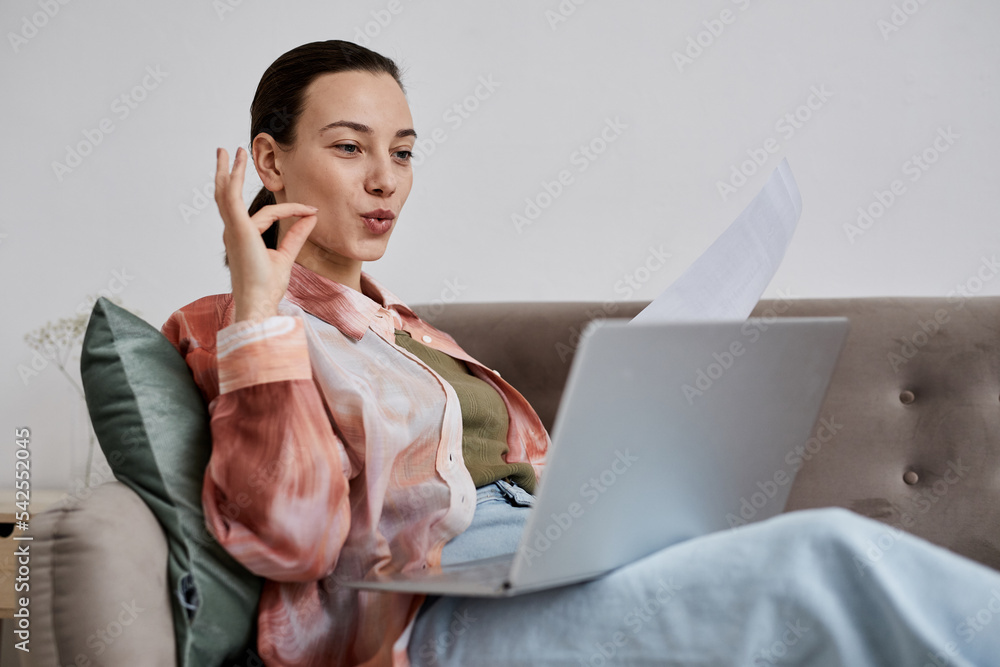 Young confident tutor with paper sitting on couch with laptop on her knees and explaining pronounciation points to online student - obrazy, fototapety, plakaty 