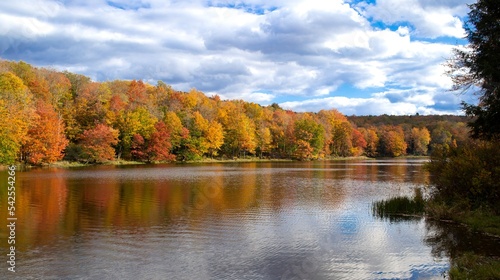 Calm lake surrounded by forest with Autumn leaf color, reflections of the trees in the water, blue sky with clouds, New York State, USA