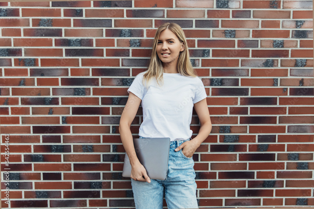 Photo of a beautiful modern woman standing on a brick exterior wall holding a silver laptop in her hands