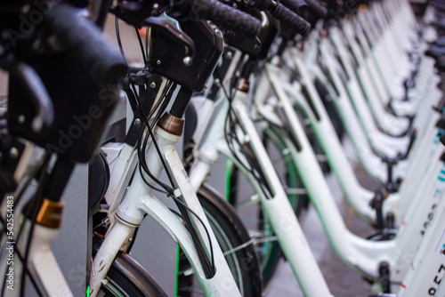 Lots of white bicycles stand in a row in bicycle parking lot. Renting and sharing bikes. Eco-friendly transportation for sports, travel in a city. Green transport rent. Handlebars, wheels, bike frames