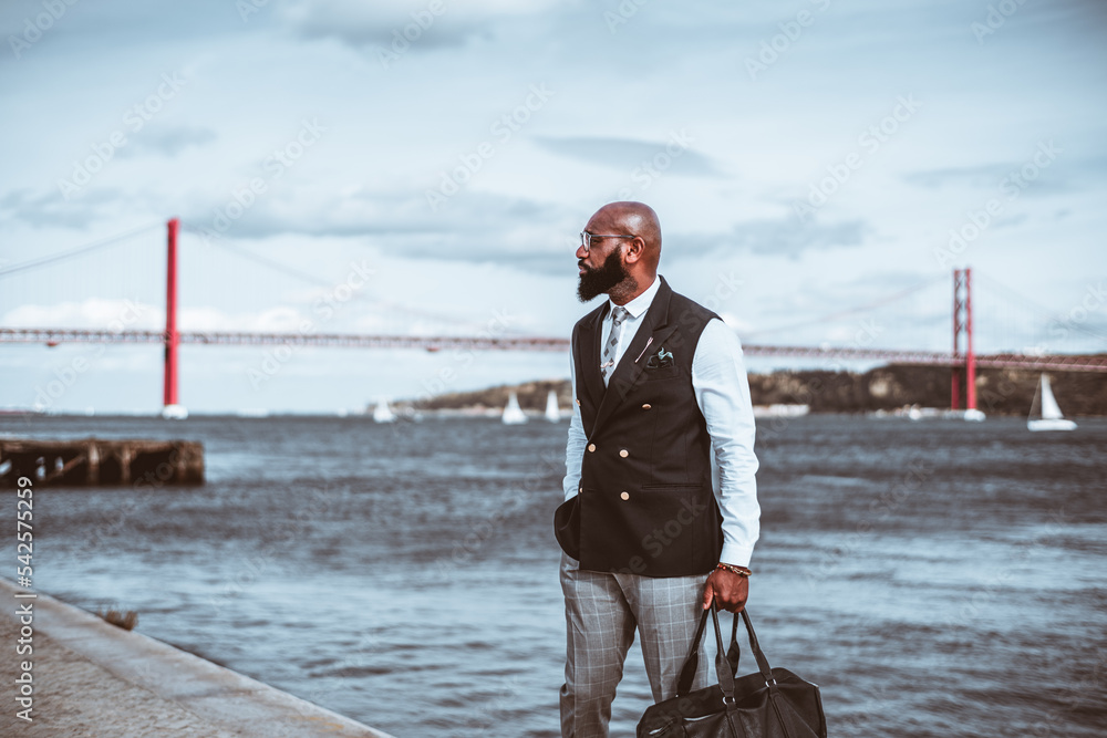 Naklejka premium An outdoor shot with a fashionable handsome mature bald black man entrepreneur with a beautiful beard, bag in his hand, eyeglasses, and a suit with a vest; a red rope bridge in a defocused background