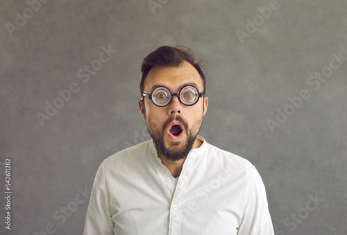 Portrait of funny shocked man round hilarious glasses standing on grey background. Puzzled bearded adult businessman staring eyes and opened mouth looking at camera. Human face expression and reaction