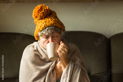 Elderly woman in ginger cap wrapped in blanket and drinking cup of tea. Old grandma feeling cold at home. Energy crisis.