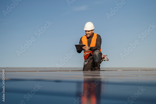 A supervisor is crouching on the roof and checking on solar panels over the tablet.